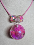 A pink cord bracelet displayed on a white fabric jewelry stand, featuring a large round clear resin bead filled with pink glitter and two smaller iridescent beads.