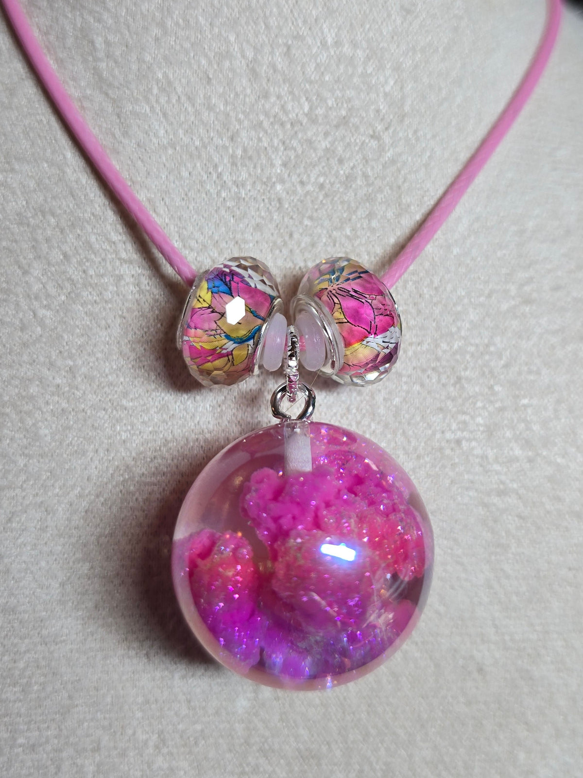 A pink cord bracelet displayed on a white fabric jewelry stand, featuring a large round clear resin bead filled with pink glitter and two smaller iridescent beads.