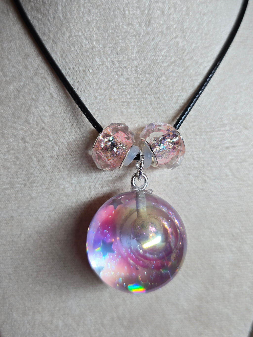A close-up of a necklace displayed on a beige jewelry bust. The necklace has a black cord with three faceted light pink beads and a large, round, iridescent resin bead pendant that reflects pink, purple, and blue colors.