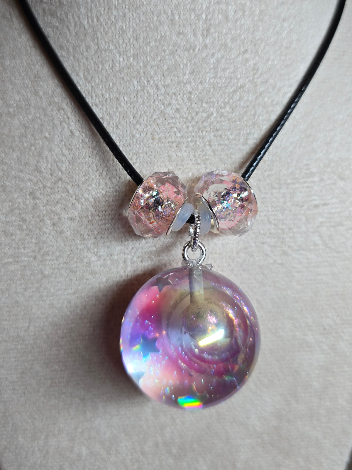 A close-up of a necklace displayed on a beige jewelry bust. The necklace has a black cord with three faceted light pink beads and a large, round, iridescent resin bead pendant that reflects pink, purple, and blue colors.