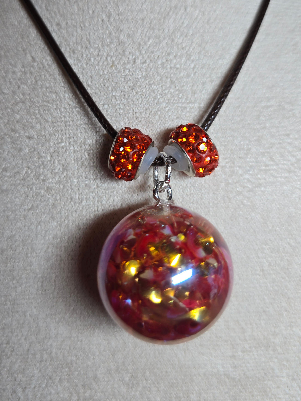 A close-up of a necklace displayed on a white jewelry bust. The necklace has a dark cord and a large round clear resin pendant filled with pink, red, and gold glitter, connected by silver-toned beads with orange rhinestones.