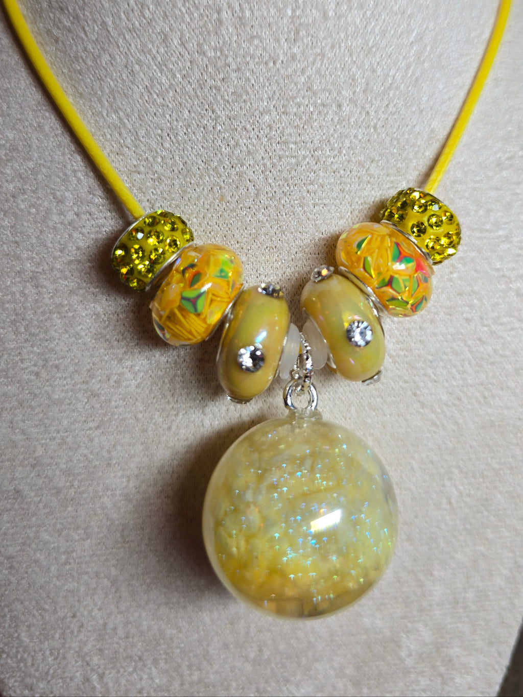 A yellow cord bracelet displayed on a beige fabric jewelry stand, featuring a large round resin translucent yellow bead with glitter, four patterned yellow glass beads with rhinestones, and two textured yellow beads.