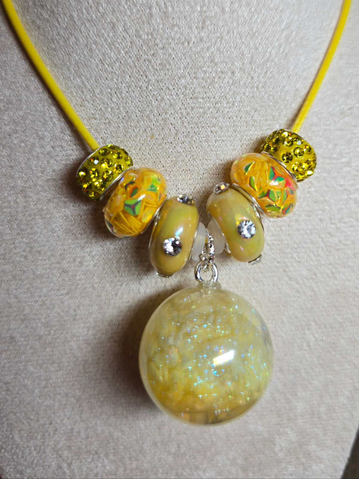 A yellow cord bracelet displayed on a beige fabric jewelry stand, featuring a large round resin translucent yellow bead with glitter, four patterned yellow glass beads with rhinestones, and two textured yellow beads.