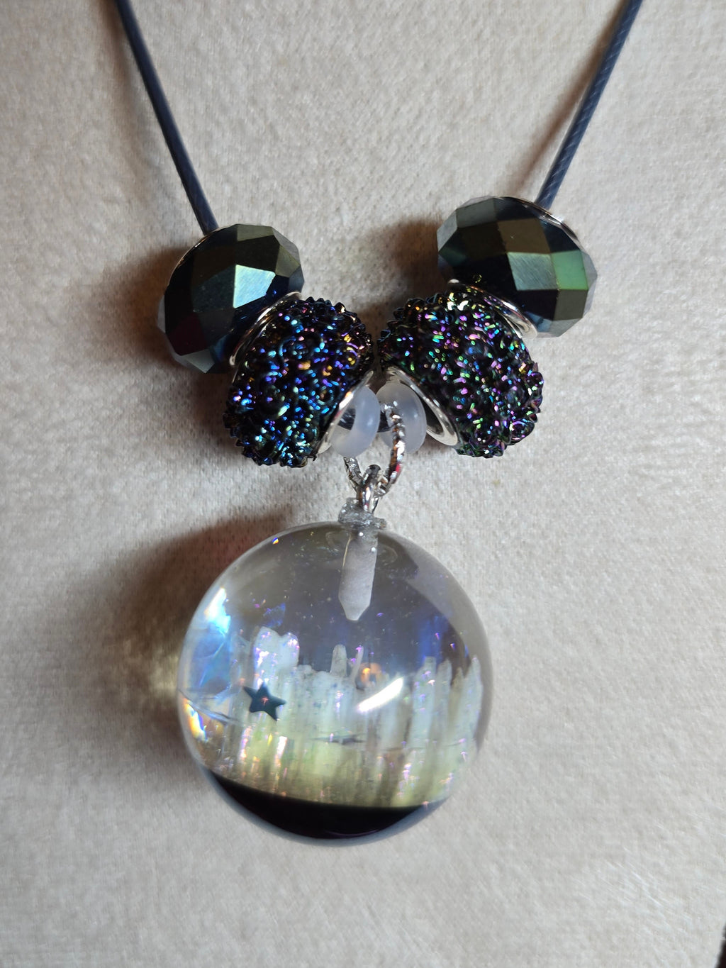 A beaded necklace displayed on a cream-colored jewelry bust. The necklace features a black cord, three faceted dark beads, two round iridescent glittery beads, and a large resin glass orb pendant containing a miniature landscape scene with a star.