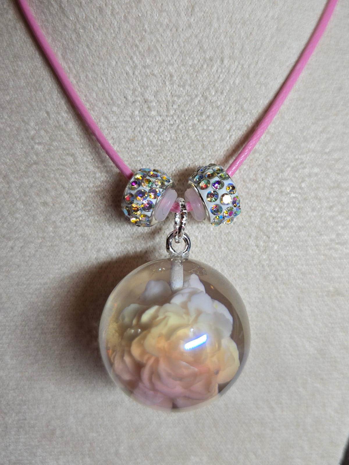 A pink cord bracelet displayed on a beige jewelry bust, featuring two silver rhinestone-covered beads and a large clear resin oval pendant containing a pale pink and yellow flower.