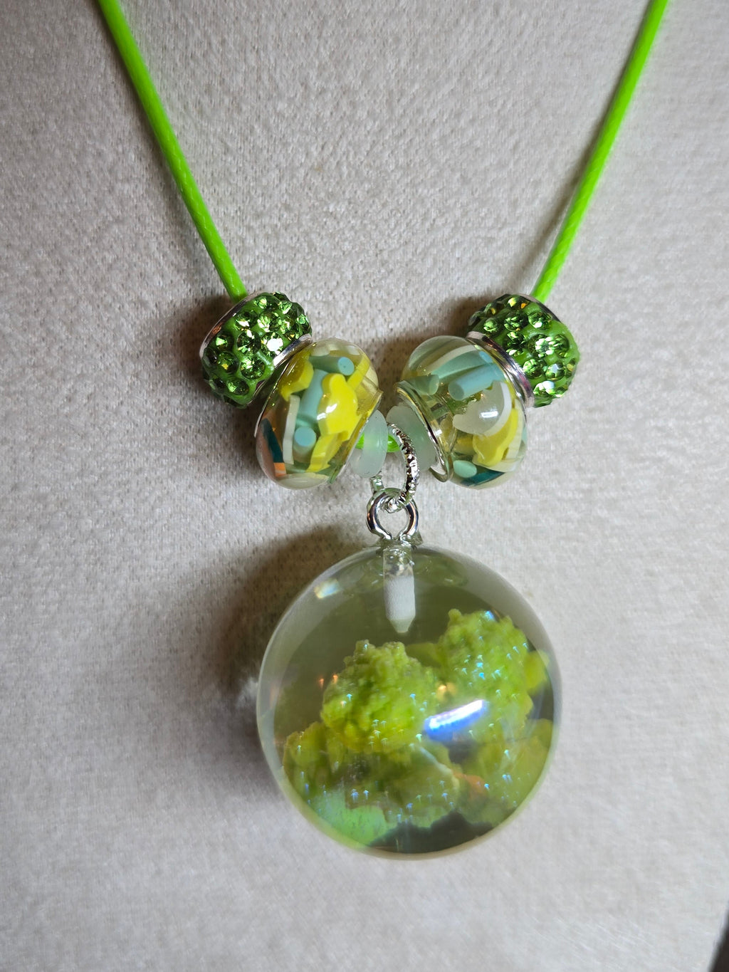 A handmade necklace with a bright neon green cord, featuring three decorative resin beads with yellow and blue swirls and two textured green beads, hanging from a silver clasp over a clear resin pendant filled with green moss or small leaves, displayed on a beige fabric mannequin bust.