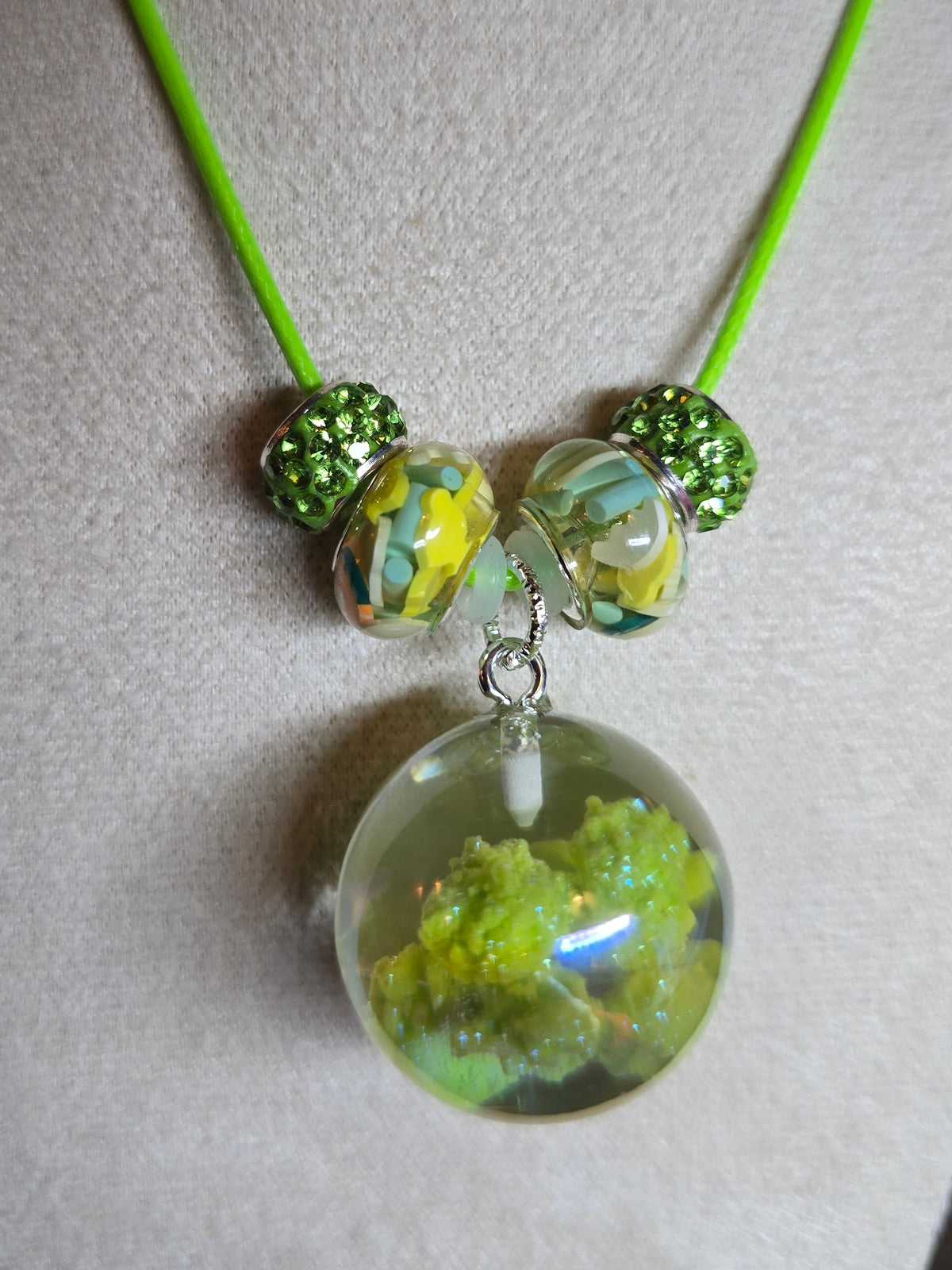 A handmade necklace with a bright neon green cord, featuring three decorative resin beads with yellow and blue swirls and two textured green beads, hanging from a silver clasp over a clear resin pendant filled with green moss or small leaves, displayed on a beige fabric mannequin bust.