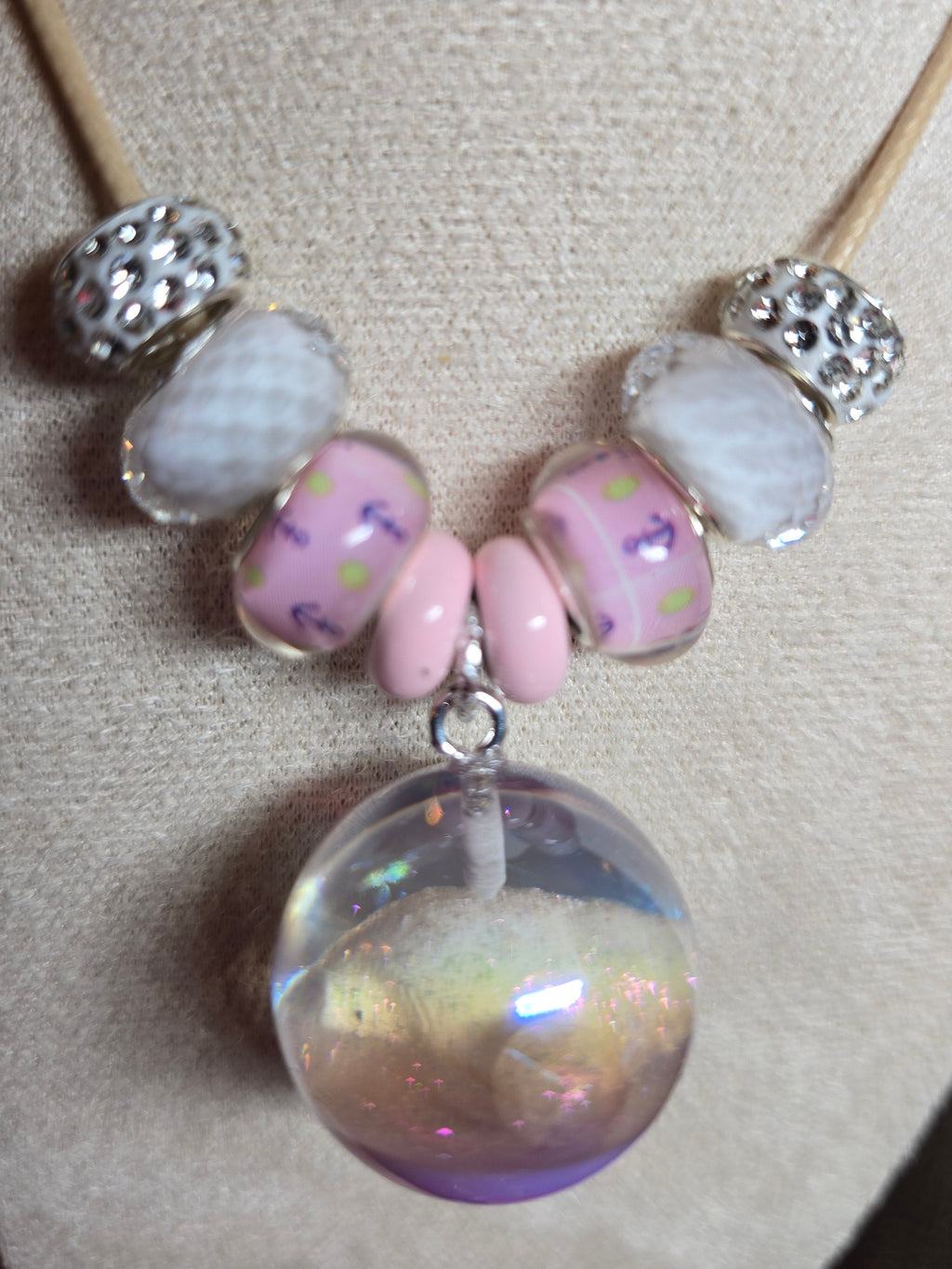 A close-up of a handmade beaded necklace displayed on a beige fabric jewelry bust. The necklace features a tan cord, silver spacer beads, light pink and white beads, and a large, round, iridescent purple resin pendant.