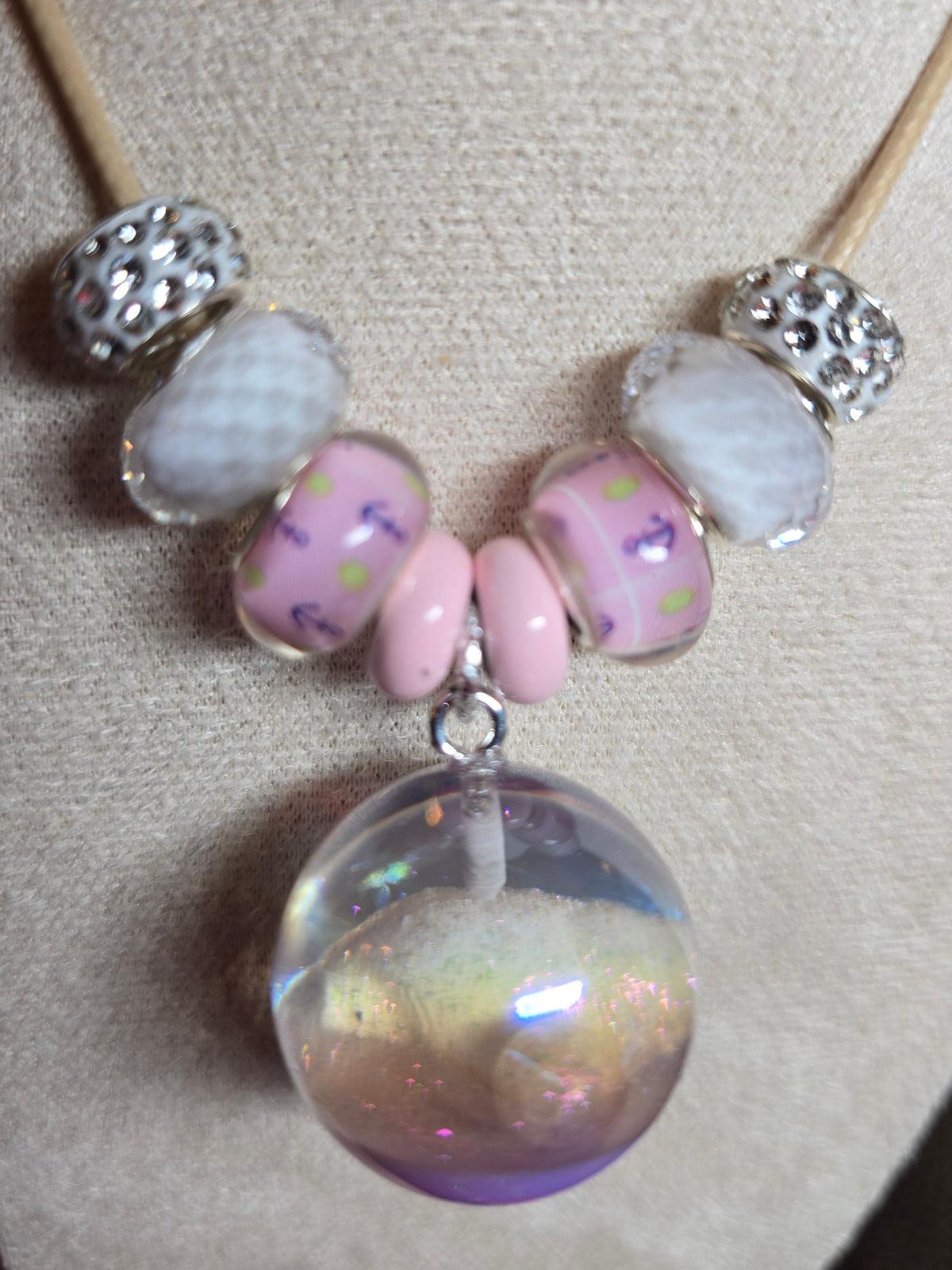 A close-up of a handmade beaded necklace displayed on a beige fabric jewelry bust. The necklace features a tan cord, silver spacer beads, light pink and white beads, and a large, round, iridescent purple resin pendant.
