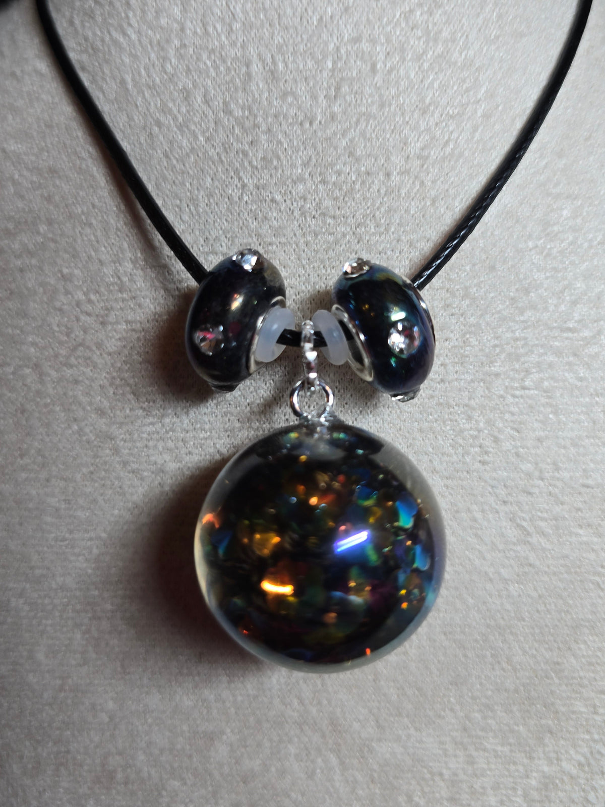 A black cord necklace with a large, glossy black resin bead filled with iridescent glitter, accented by two smaller black beads and silver spacer rings, displayed on a light gray textured surface.
