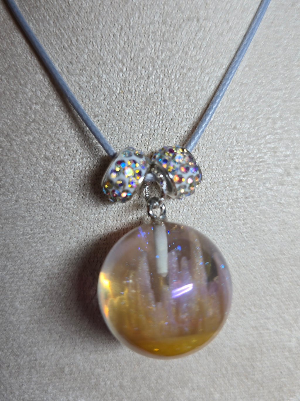 A close-up of a necklace displayed on a beige fabric jewelry bust. The necklace has a light blue cord, two silver-toned oval beads covered in iridescent rhinestones, and a large round resin glass pendant filled with gold glitter and sparkles.