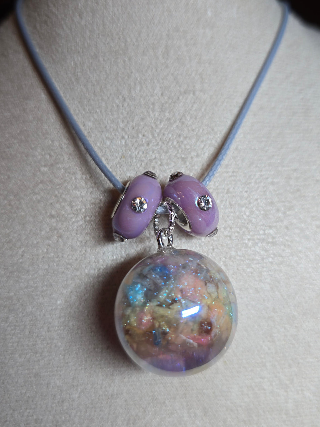 A close-up of a necklace displayed on a cream-colored jewelry bust. The necklace features a light blue cord, two lavender purple beads with small silver rhinestone accents, and a large clear resin orb filled with multicolored glitter.