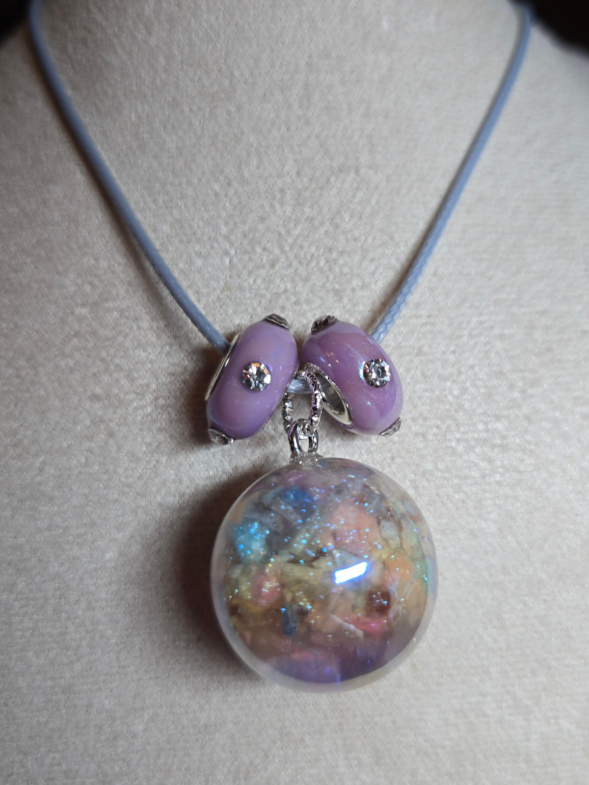 A close-up of a necklace displayed on a cream-colored jewelry bust. The necklace features a light blue cord, two lavender purple beads with small silver rhinestone accents, and a large clear resin orb filled with multicolored glitter.