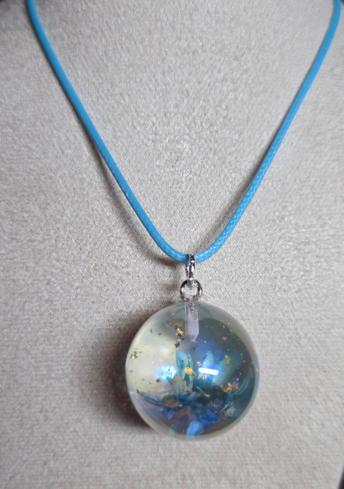 A light blue cord necklace with a clear spherical pendant containing iridescent blue, gold, and white inclusions, displayed on a beige fabric jewelry stand.