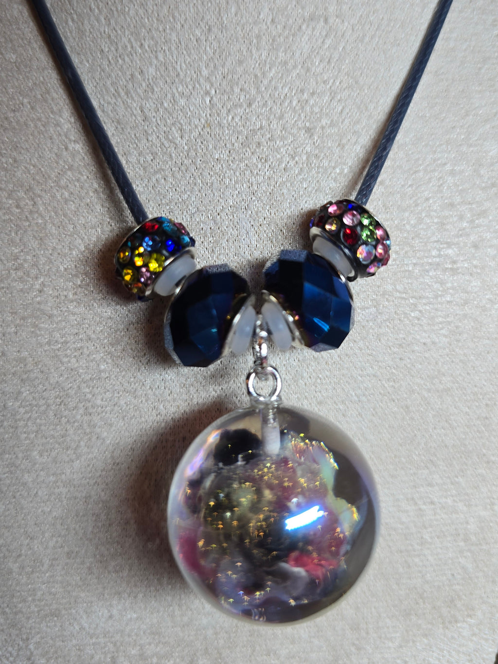 A beaded necklace displayed on a beige fabric jewelry bust. The necklace features a dark cord, two silver-toned beads with iridescent multicolor crystals, two dark blue faceted beads, and a large clear resin orb pendant filled with gold glitter and colorful inclusions.