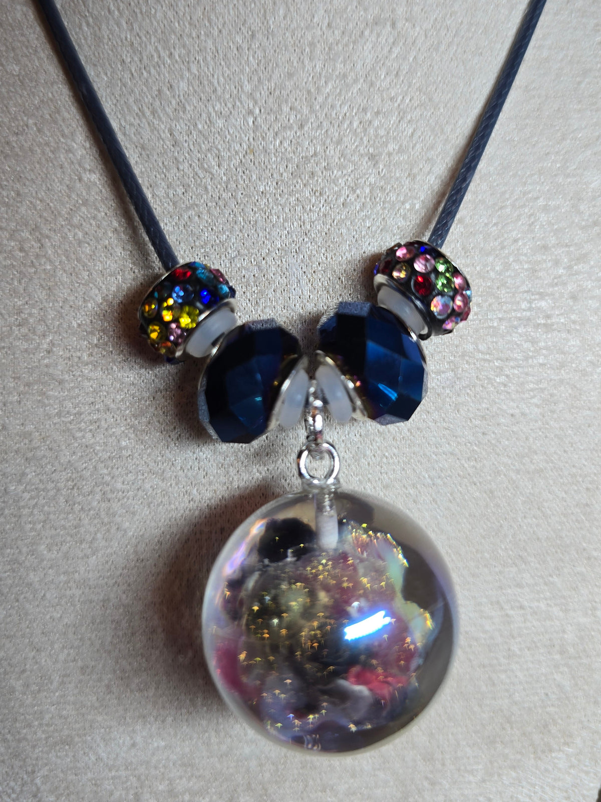 A beaded necklace displayed on a beige fabric jewelry bust. The necklace features a dark cord, two silver-toned beads with iridescent multicolor crystals, two dark blue faceted beads, and a large clear resin orb pendant filled with gold glitter and colorful inclusions.