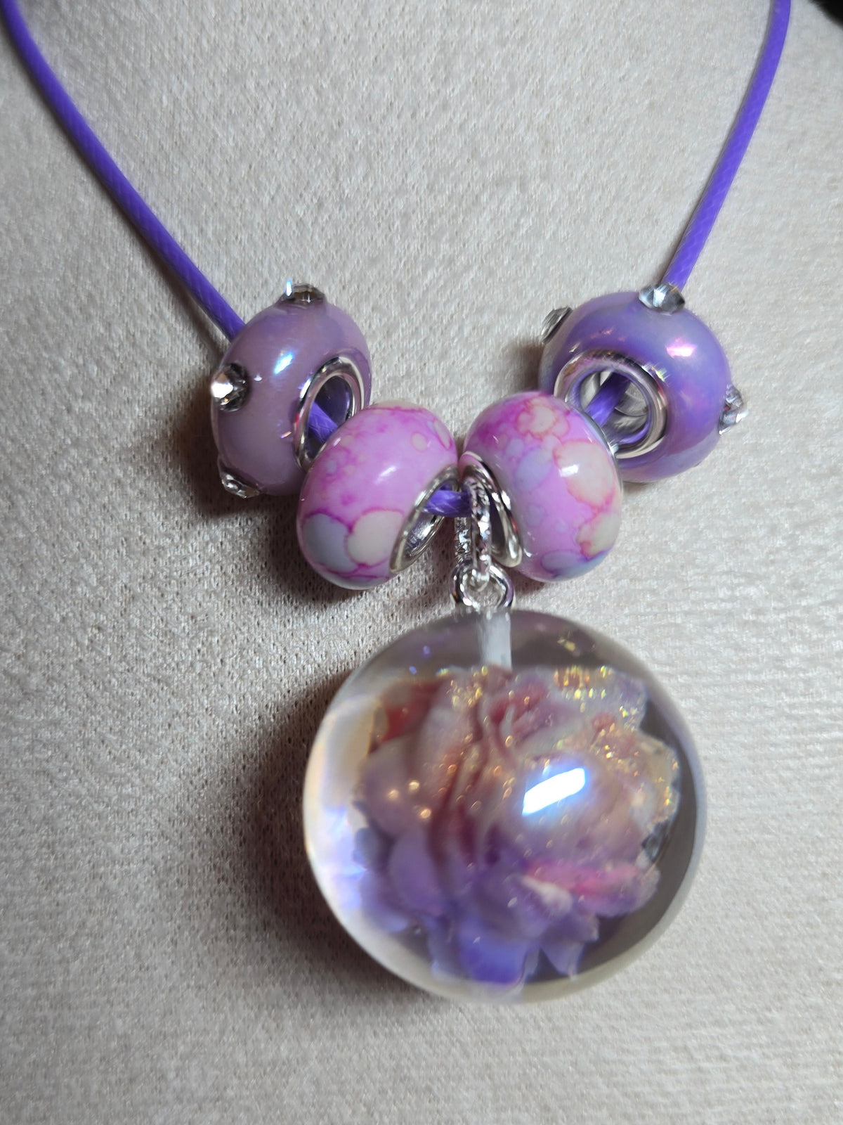 A close-up of a jewelry charm or bead component on a light fabric surface. The product includes a purple cord, lavender and pink marbled beads, silver-toned metal rings, and a large clear resin round bead containing a purple and iridescent floral design.