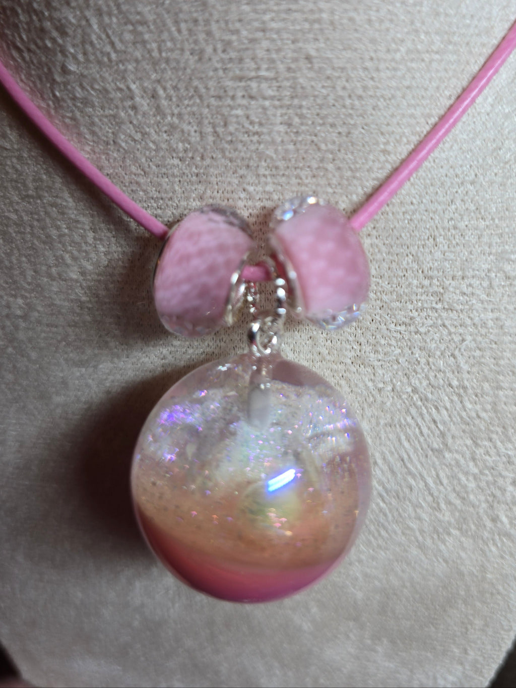 A pink cord bracelet displayed on a beige fabric jewelry stand, featuring a large iridescent pink resin bead and two smaller pink heart-shaped beads separated by a silver spacer.