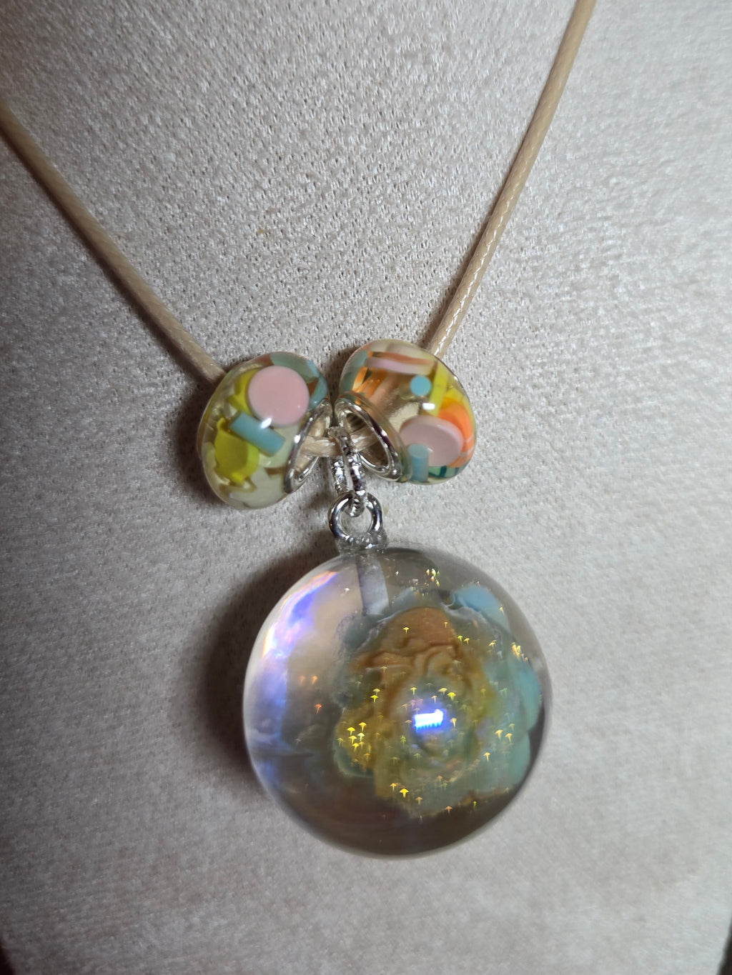 A close-up of a necklace displayed on a cream fabric jewelry bust. The necklace has a thin tan cord and a large clear resin round bead filled with iridescent glitter, with two smaller marbled beads in pastel colors below.