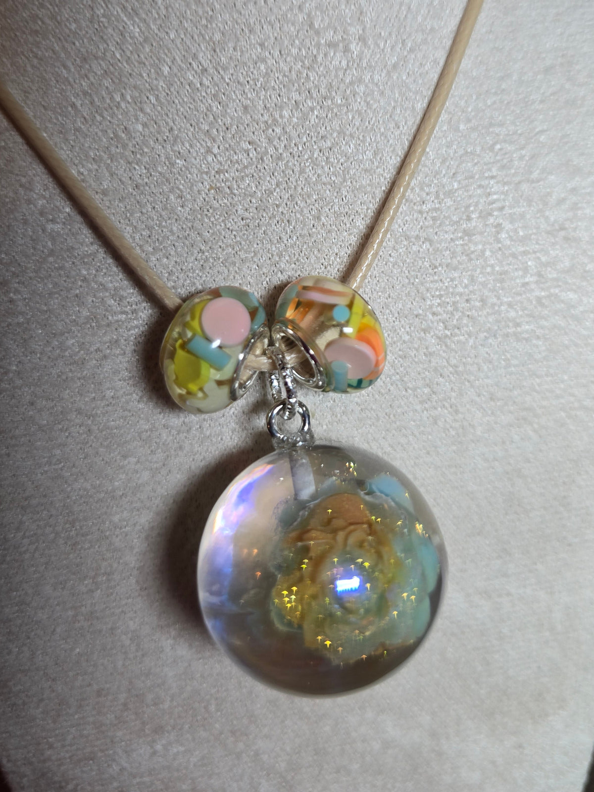 A close-up of a necklace displayed on a cream fabric jewelry bust. The necklace has a thin tan cord and a large clear resin round bead filled with iridescent glitter, with two smaller marbled beads in pastel colors below.