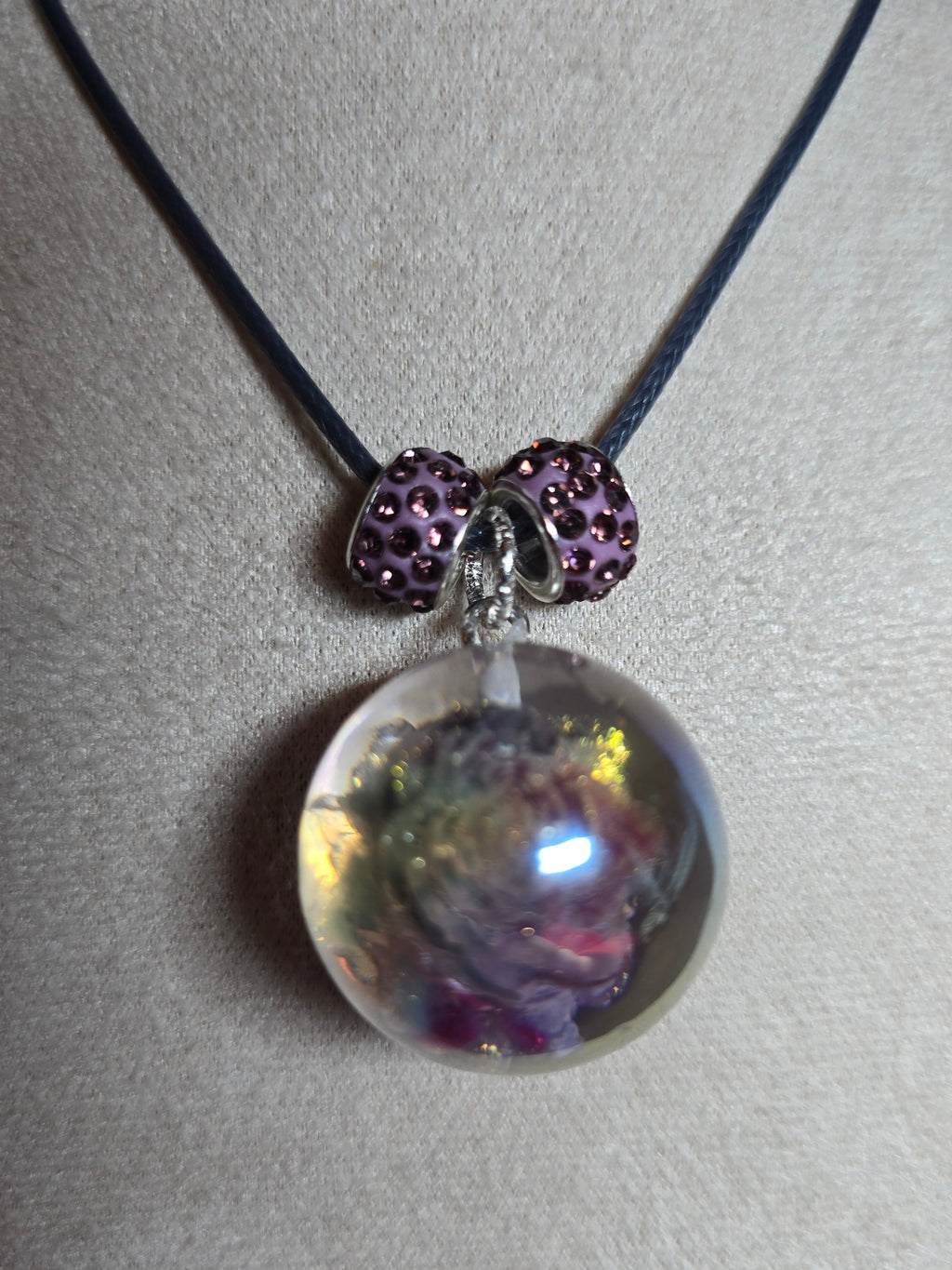 A necklace displayed on a beige fabric jewelry bust, featuring a dark cord, two purple rhinestone-covered beads, and a large clear resin orb pendant containing dried floral elements.