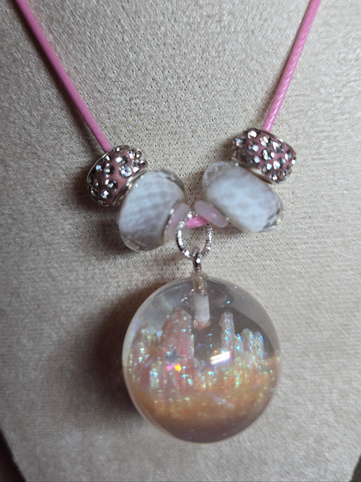 A close-up of a jewelry charm or resin bead assembly on a pink cord, featuring a large iridescent clear sphere, two white marbled beads, and two silver rhinestone spacers, displayed on a textured beige surface.