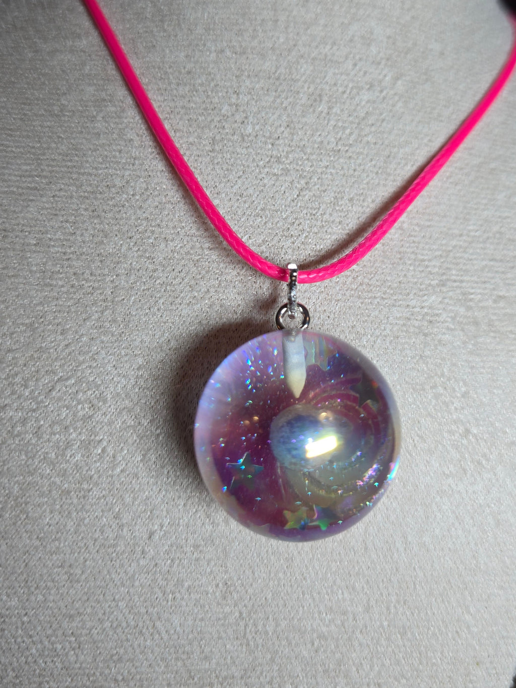 A close-up of a necklace displayed on a light gray fabric jewelry bust. The necklace has a bright pink cord and a large, round, iridescent resin bead pendant with purple, blue, and pink internal swirls.