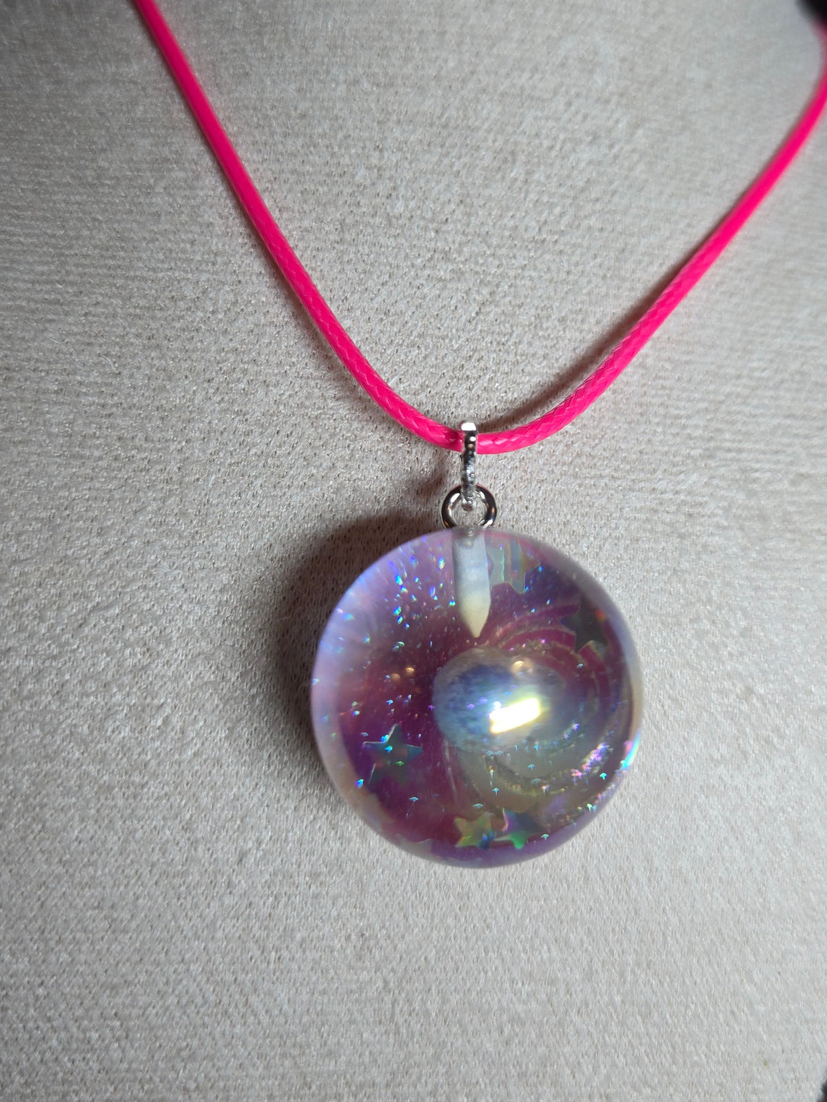 A close-up of a necklace displayed on a light gray fabric jewelry bust. The necklace has a bright pink cord and a large, round, iridescent resin bead pendant with purple, blue, and pink internal swirls.