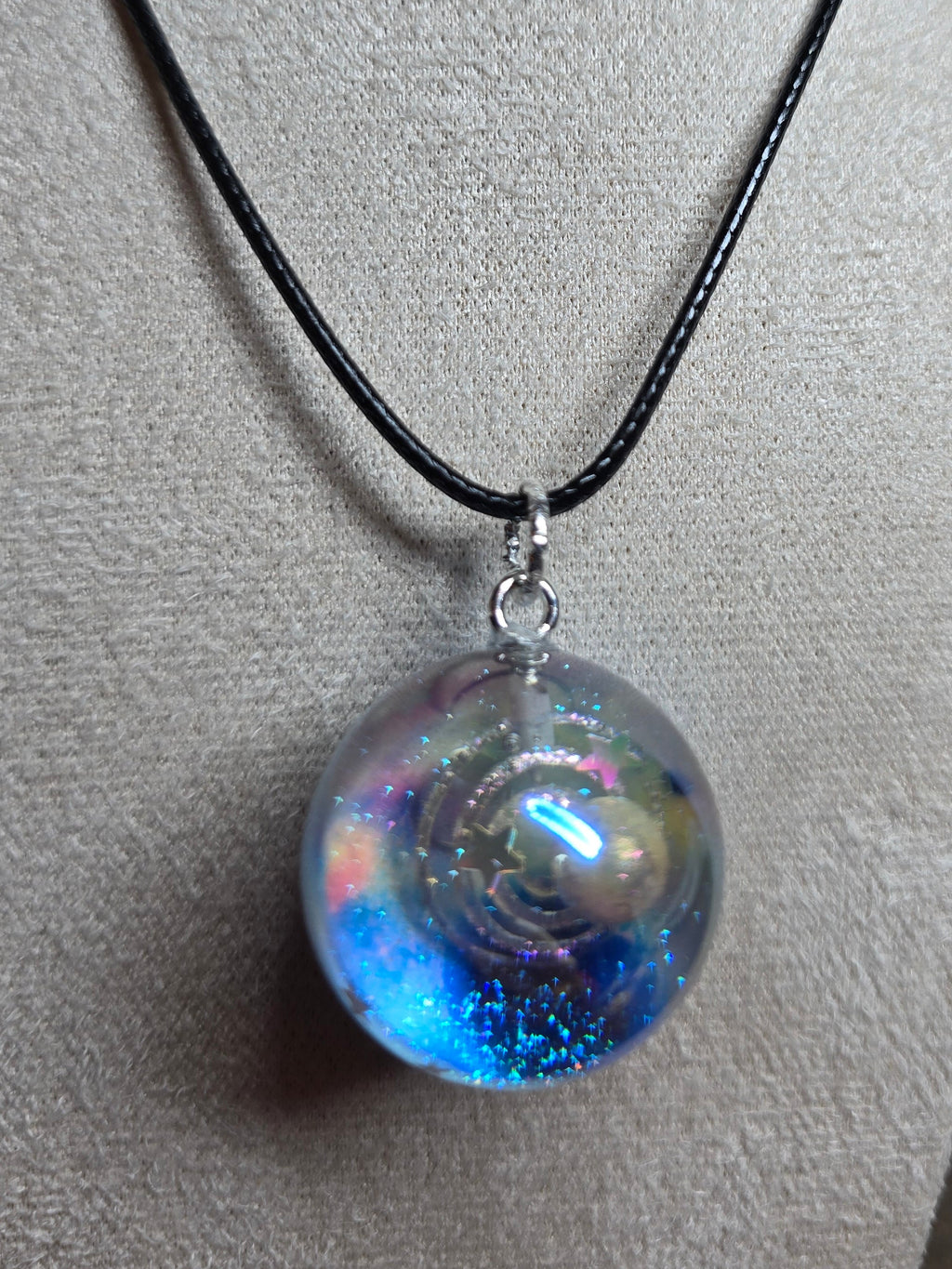 A round iridescent resin pendant necklace with blue and pink glitter, suspended on a black cord from a beige fabric mannequin bust.