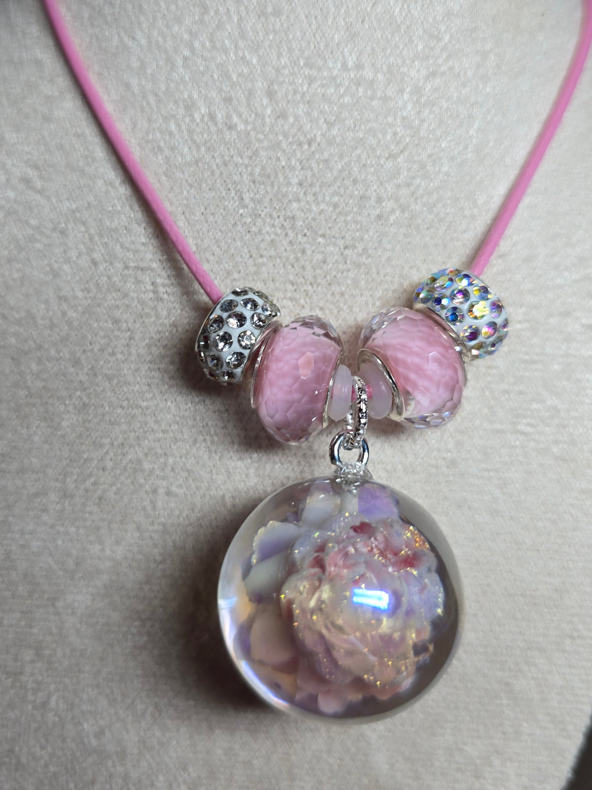 A delicate pink cord bracelet displayed on a beige fabric jewelry stand. The bracelet features three pink seashell beads, two silver-toned rhinestone-encrusted spacer beads, and a large clear resin round dome pendant containing dried pink and white flower petals.