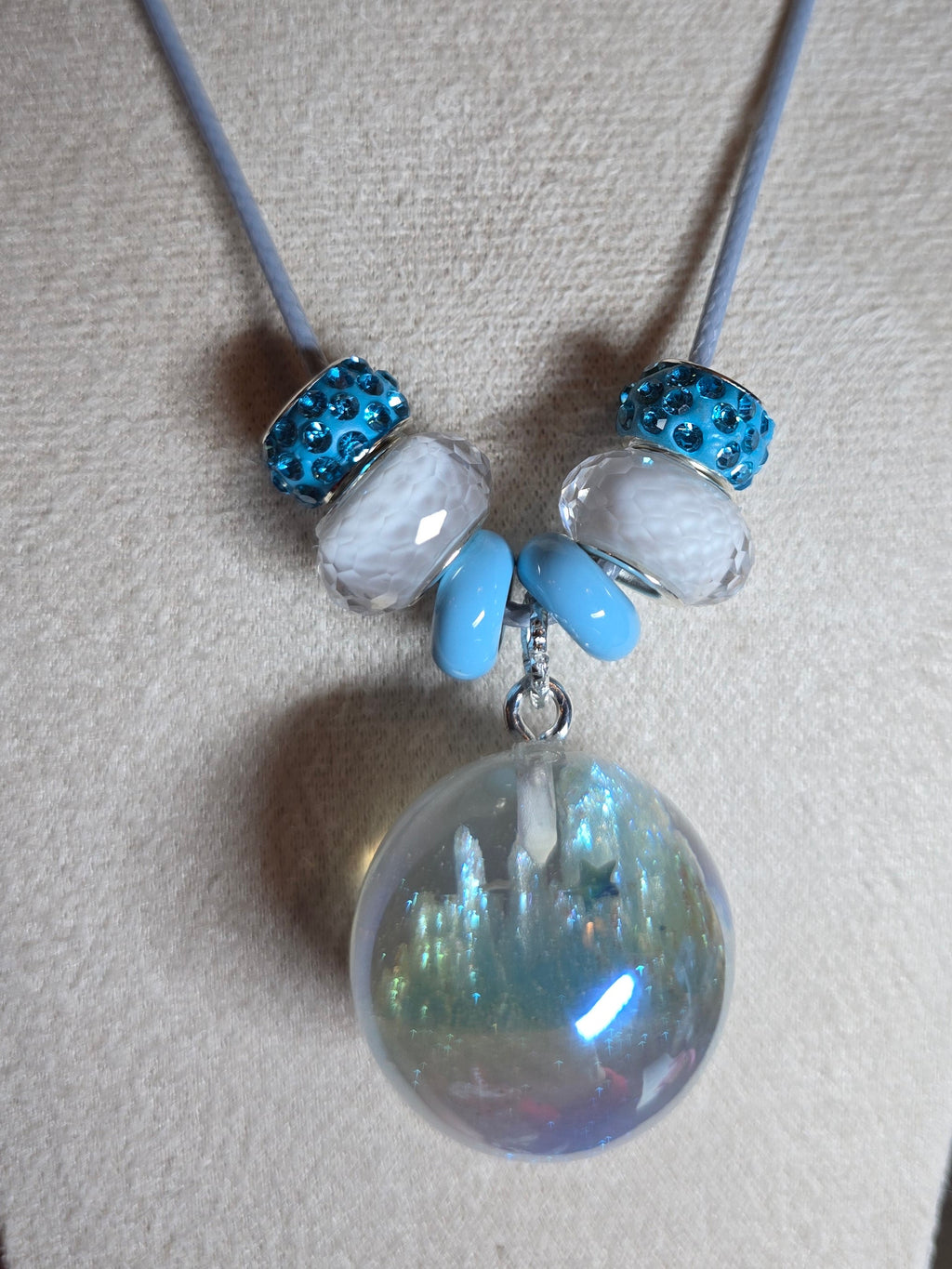 A beaded necklace displayed on a beige fabric jewelry bust. The necklace has a light gray cord, two textured blue beads, two milky white beads, two light blue beads, and a large iridescent resin sphere pendant.