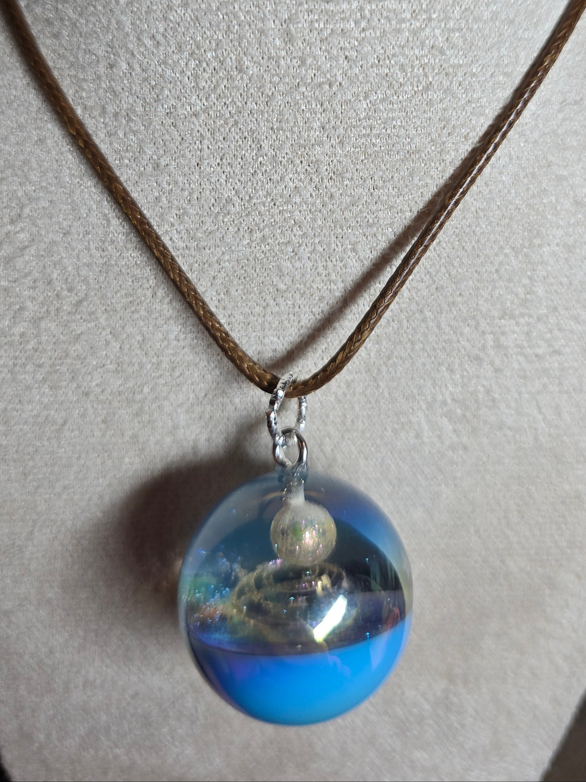 A brown cord necklace with a clear resin spherical pendant containing blue sand and a small white pearl, displayed on a beige fabric mannequin bust.