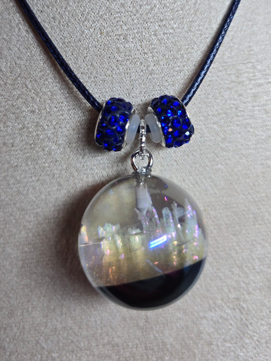 A necklace displayed on a beige fabric mannequin bust, featuring a black cord, two dark blue beaded accents, and a large clear resin orb pendant containing a dark bottom layer and iridescent upper layer.