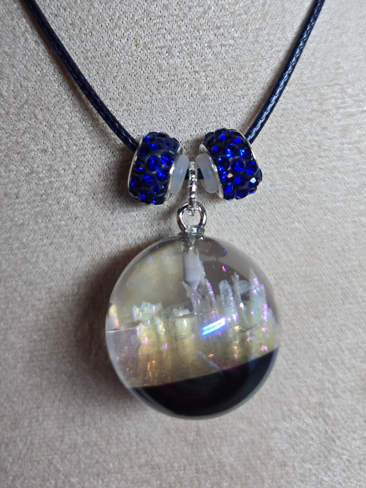 A necklace displayed on a beige fabric mannequin bust, featuring a black cord, two dark blue beaded accents, and a large clear resin orb pendant containing a dark bottom layer and iridescent upper layer.